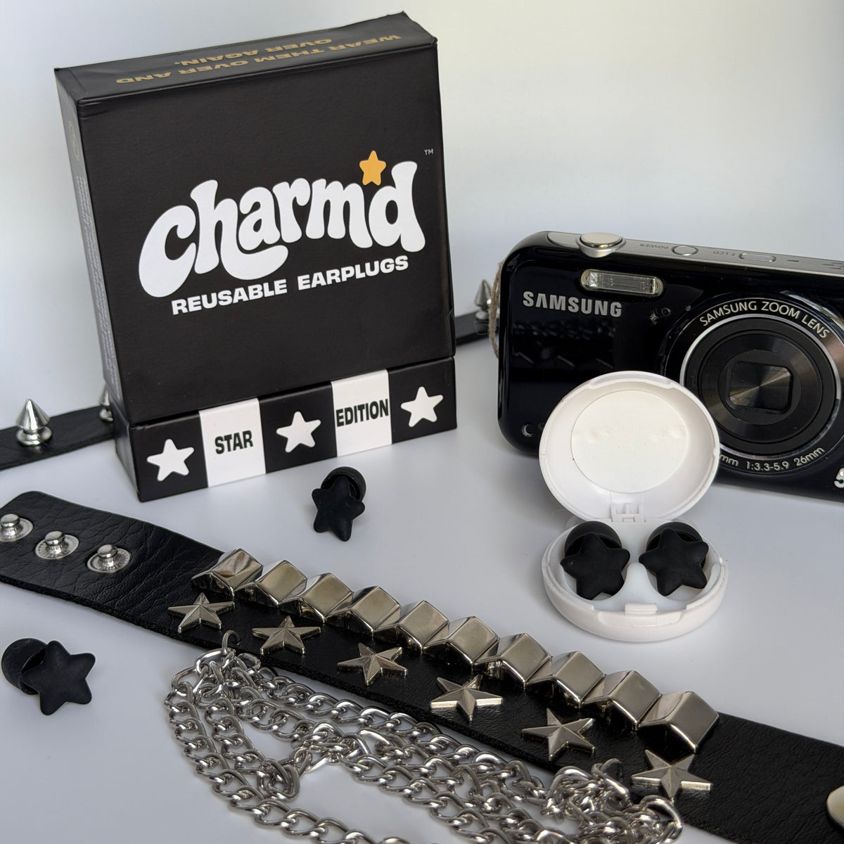 Charmd reusable earplugs set displayed with a black branded box, a black Samsung camera, a white earplug case, and a black studded leather strap with silver chains and star-shaped earplugs on a white background.