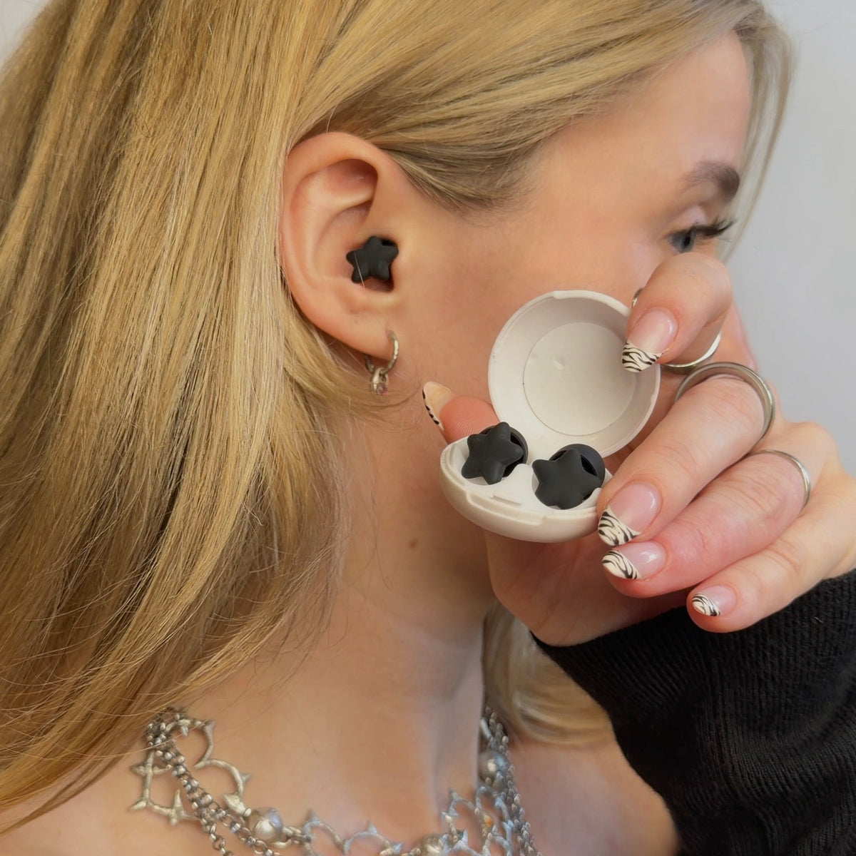 A close-up lifestyle shot of a woman with blonde hair wearing black star-shaped stud earrings and holding a small white earbud charging case next to her ear.