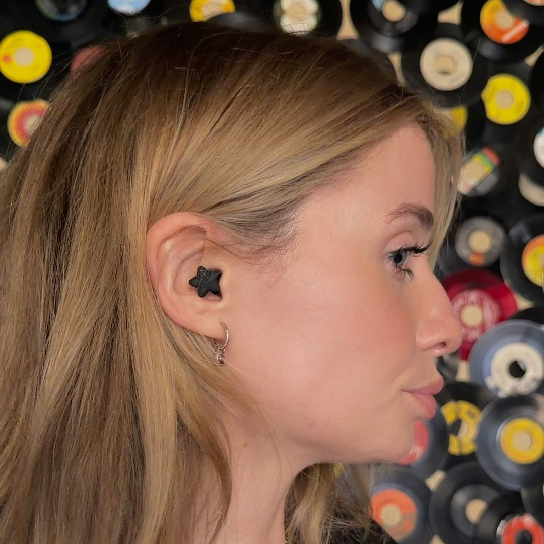 A woman in profile wearing a black star-shaped stud earring and a small silver hoop earring, with layered silver necklaces, against a background of colorful vinyl records.